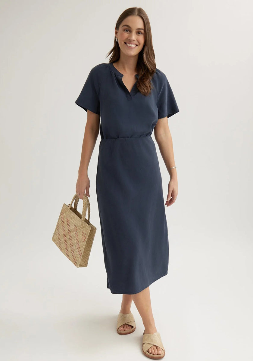 A woman with long brown hair wears a navy blue Bella Dahl Lyocell midi skirt with a cinched waist. She holds a woven tan handbag, wears beige slide sandals, and smiles while standing against a plain light background.
