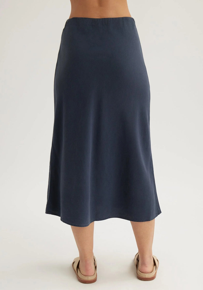 A person is shown from the waist down, wearing a Bella Dahl high-waisted, navy blue Lyocell midi skirt with a slight flare. They stand barefoot except for beige wide-strap sandals against a plain, light-colored background.