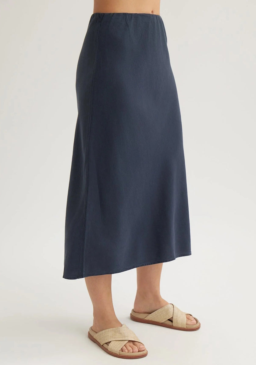On a light background, only the lower torso and legs of a person are visible, wearing a dunkelblauer Bella Dahl Lyocell-Midirock mit elastischem Bund und beigeflochtenen Sandalen mit Kreuzriemen.