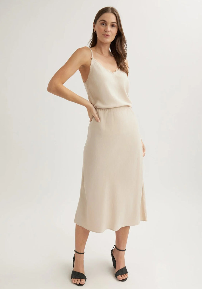 A woman with long brown hair models a sleeveless, light beige Bella Dahl Lyocell midi slip dress with thin straps, an A-line skirt, and a subtle v-neckline against a plain background. She pairs it with black ankle-strap high-heeled sandals.