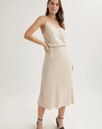 A woman with long brown hair models a sleeveless, light beige Bella Dahl Lyocell midi slip dress with thin straps, an A-line skirt, and a subtle v-neckline against a plain background. She pairs it with black ankle-strap high-heeled sandals.