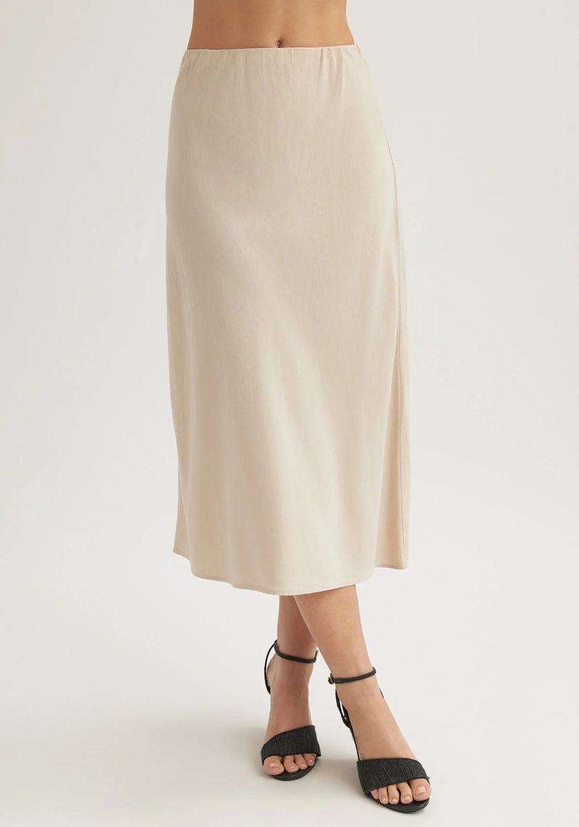A person wears a high-waisted, light beige Lyocell midi flared skirt by Bella Dahl with black open-toe ankle strap heels. The waist-down photo is set against a plain white background.