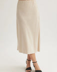 A person wears a high-waisted, light beige Lyocell midi flared skirt by Bella Dahl with black open-toe ankle strap heels. The waist-down photo is set against a plain white background.