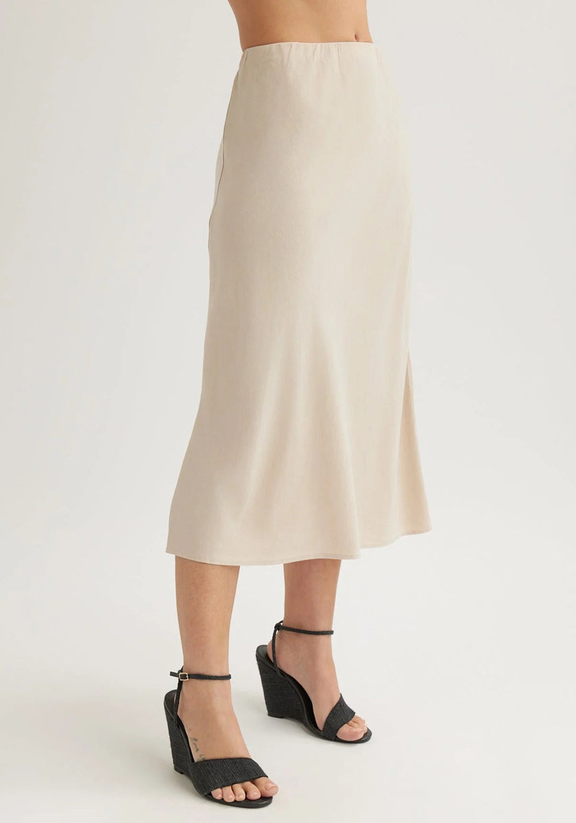 Wearing a Bella Dahl high-waisted, light beige Lyocell midi skirt with a slight flare, paired with black wedge ankle-strap sandals. The plain white background emphasizes the skirt. Upper body is not visible.