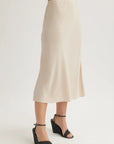 Wearing a Bella Dahl high-waisted, light beige Lyocell midi skirt with a slight flare, paired with black wedge ankle-strap sandals. The plain white background emphasizes the skirt. Upper body is not visible.