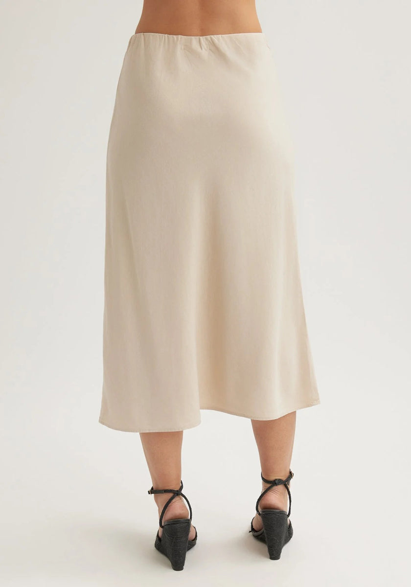 A person faces away, wearing a cream-colored Bella Dahl Lyocell midi skirt with a slight flare. The lightweight, silky skirt is styled with black strappy low-heeled sandals against a plain white background.