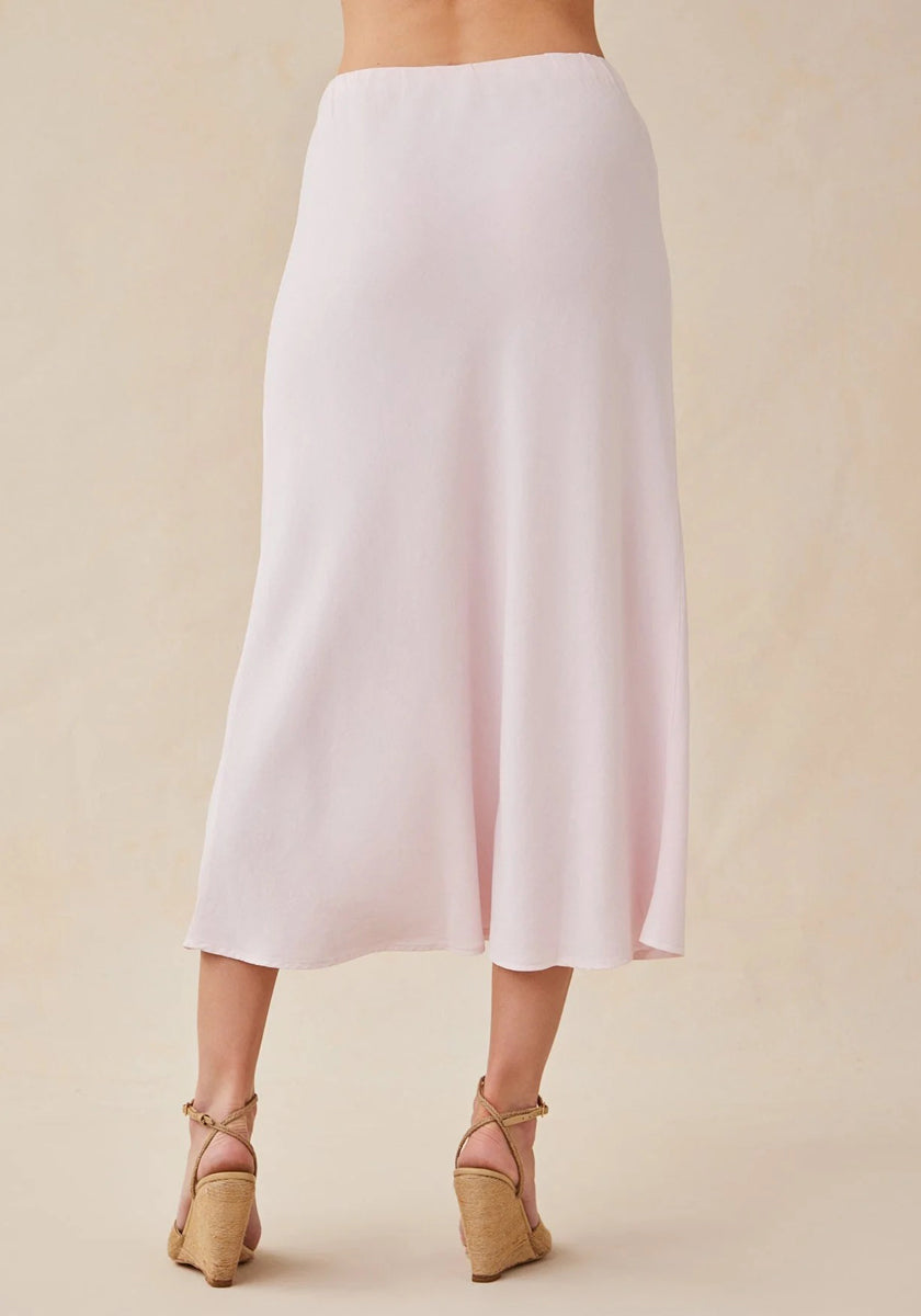 A person faces away from the camera in a Bella Dahl light pink Lyocell midi skirt with a soft drape to mid-calf. They wear tan strappy wedge sandals. The neutral beige background and top half out of frame draw focus to the skirt.