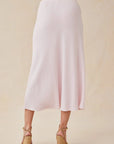 A person faces away from the camera in a Bella Dahl light pink Lyocell midi skirt with a soft drape to mid-calf. They wear tan strappy wedge sandals. The neutral beige background and top half out of frame draw focus to the skirt.
