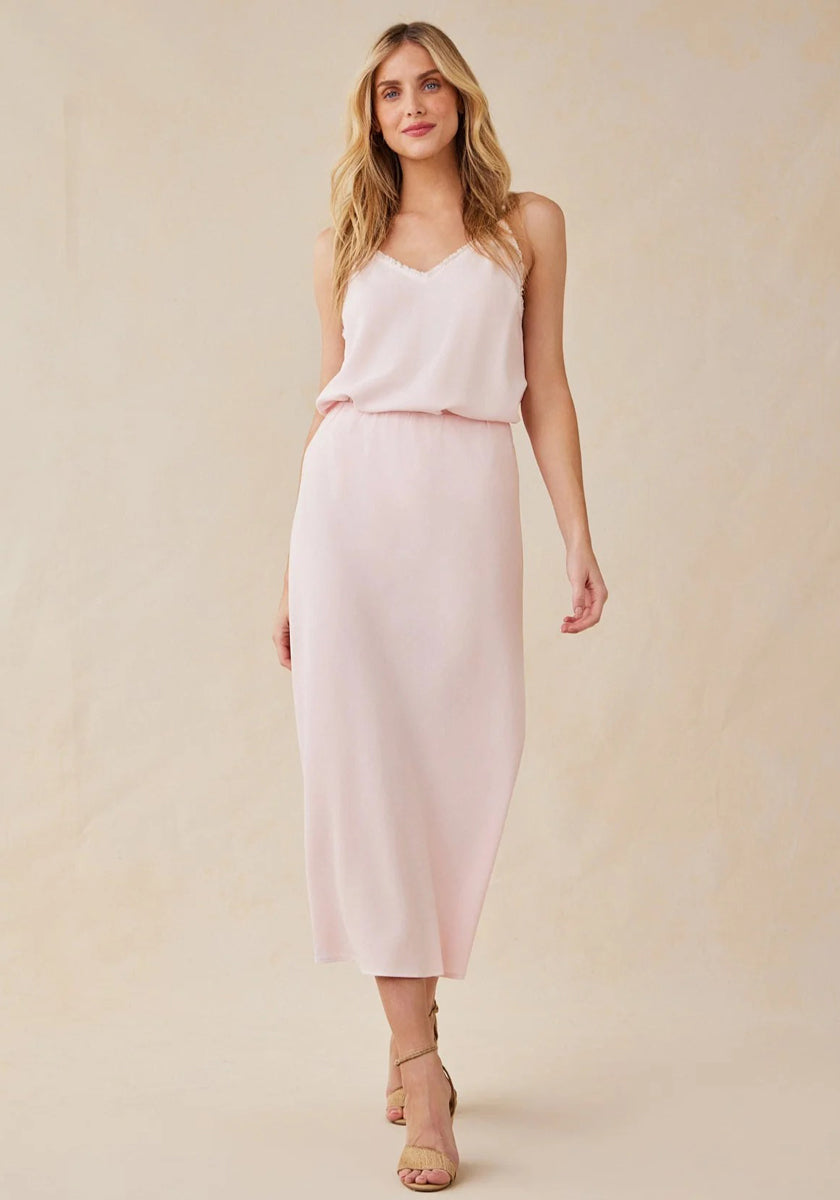 A woman with long, wavy blonde hair stands against a neutral background in a light pink Bella Dahl Lyocell midi dress with flared skirt and subtle V-neck, wearing beige strappy heels and smiling gently with one relaxed hand at her side.