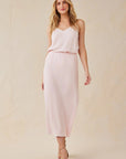 A woman with long, wavy blonde hair stands against a neutral background in a light pink Bella Dahl Lyocell midi dress with flared skirt and subtle V-neck, wearing beige strappy heels and smiling gently with one relaxed hand at her side.