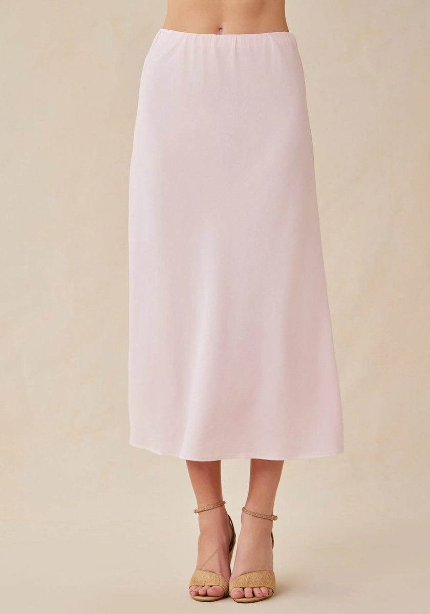 A person stands against a beige background, wearing a pale pink Bella Dahl midi-length A-line Lyocell skirt and tan open-toe ankle-strap sandals. Only the body from waist to feet is shown.