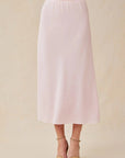 A person stands against a beige background, wearing a pale pink Bella Dahl midi-length A-line Lyocell skirt and tan open-toe ankle-strap sandals. Only the body from waist to feet is shown.