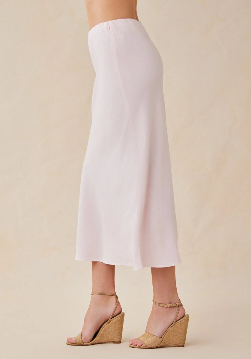 A person stands sideways in a zartrosa, midi-langer ausgestellter Rock von Bella Dahl, kombiniert mit beigen Wedge-Sandalen mit Knöchelriemen. Der Hintergrund ist schlicht und hellbeige; der Fokus liegt auf dem Outfit.