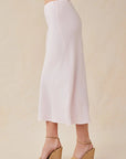 A person stands sideways in a zartrosa, midi-langer ausgestellter Rock von Bella Dahl, kombiniert mit beigen Wedge-Sandalen mit Knöchelriemen. Der Hintergrund ist schlicht und hellbeige; der Fokus liegt auf dem Outfit.