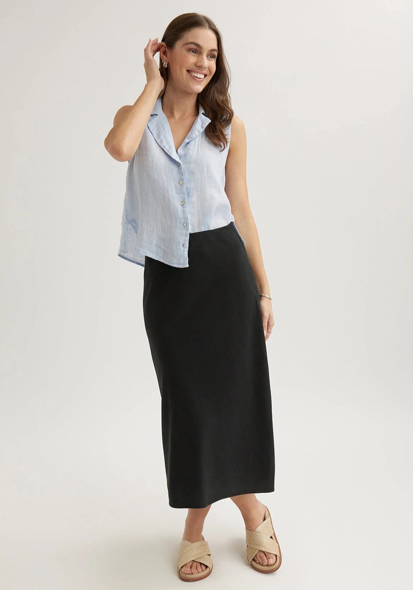 A woman with long brown hair smiles against a plain background, wearing a sleeveless light blue Bella Dahl button-up blouse and a midi flared skirt. She pairs the look with tan sandals, minimal jewelry, and touches her hair.