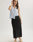 A woman with long brown hair smiles against a plain background, wearing a sleeveless light blue Bella Dahl button-up blouse and a midi flared skirt. She pairs the look with tan sandals, minimal jewelry, and touches her hair.