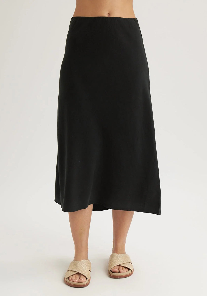 Standing against a plain, light background, a person showcases Bella Dahl’s ausgestellter Lyocell-Midi-Rock and beige crisscross sandals. The cropped image focuses on the minimalist style, with only the lower body visible.