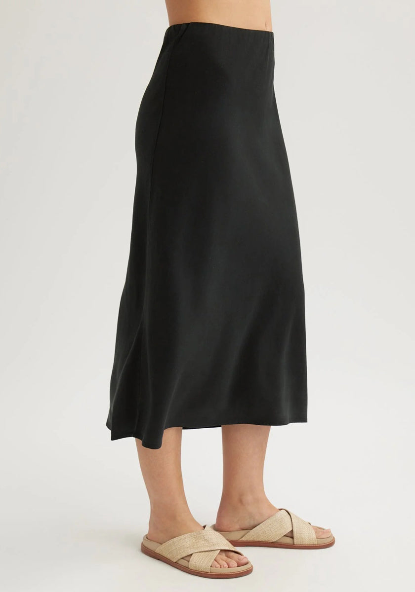 A person stands against a plain background wearing a Bella Dahl black Lyocell flared midi skirt with a slight sheen and beige woven flat sandals. The upper body is not shown.