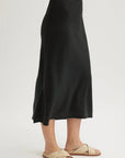 A person stands against a plain background wearing a Bella Dahl black Lyocell flared midi skirt with a slight sheen and beige woven flat sandals. The upper body is not shown.