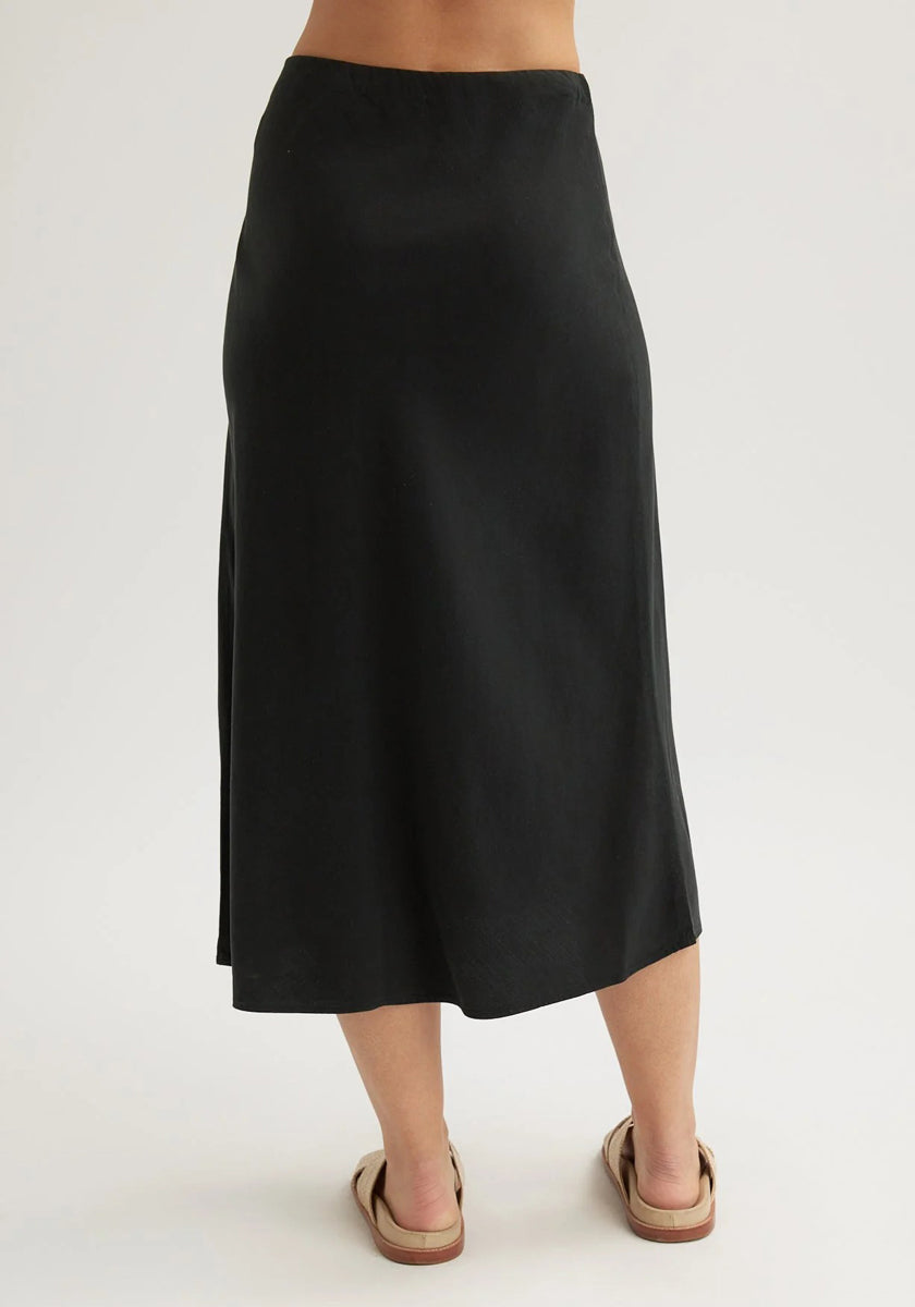 A person stands barefoot on a light surface, facing away from the camera, wearing a long black Bella Dahl Lyocell midi-length flared skirt that falls to mid-calf and tan sandals. The background is plain and light-colored.