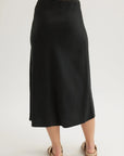 A person stands barefoot on a light surface, facing away from the camera, wearing a long black Bella Dahl Lyocell midi-length flared skirt that falls to mid-calf and tan sandals. The background is plain and light-colored.