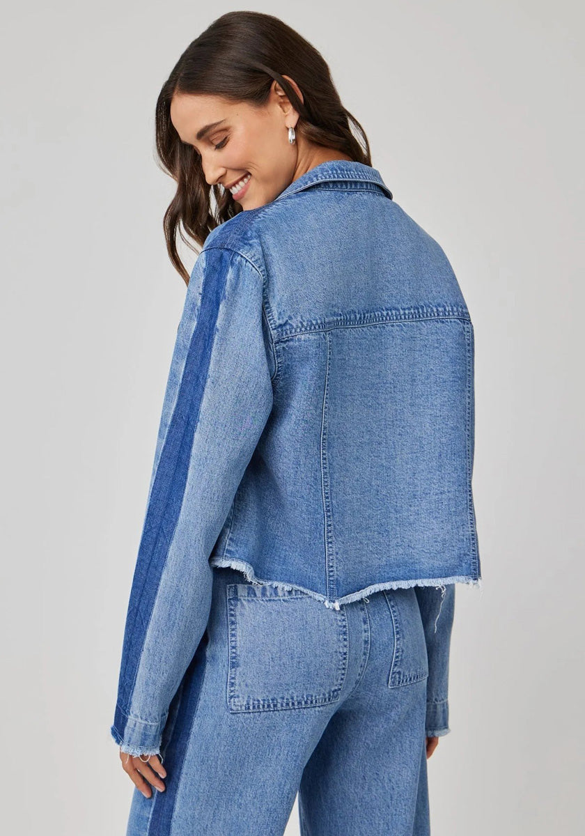 A woman with long brown hair smiles, glancing over her shoulder in a light blue denim-look cropped Bella Dahl jacket with raw, frayed hems, dark blue sleeve stripe on light gray, and matching high-waisted jeans.
