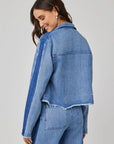 A woman with long brown hair smiles, glancing over her shoulder in a light blue denim-look cropped Bella Dahl jacket with raw, frayed hems, dark blue sleeve stripe on light gray, and matching high-waisted jeans.