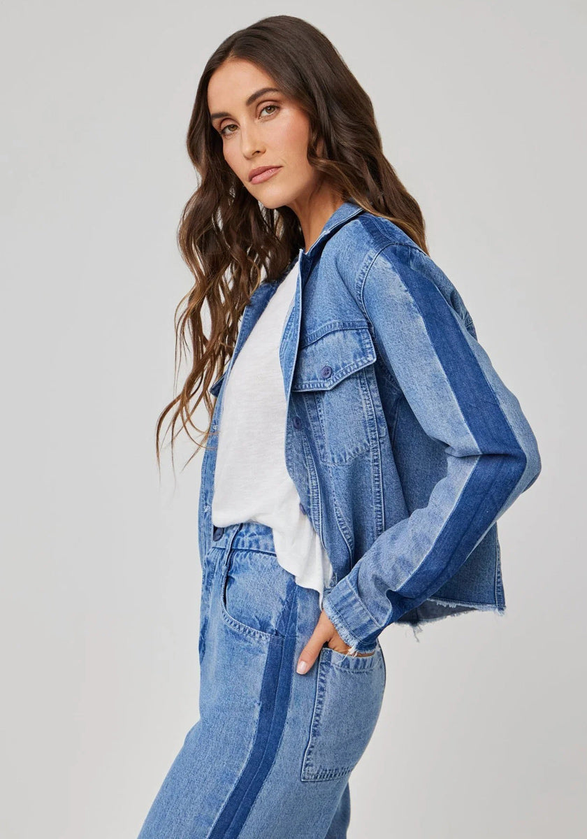 A woman with long wavy brown hair wears a light blue Bella Dahl Lyocell denim jacket and matching jeans with dark blue side stripes, standing in profile against a light gray background and looking at the camera neutrally.