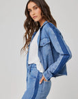 A woman with long wavy brown hair wears a light blue Bella Dahl Lyocell denim jacket and matching jeans with dark blue side stripes, standing in profile against a light gray background and looking at the camera neutrally.