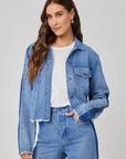 A woman with long brown hair wears a cropped Bella Dahl denim-look jacket over a white t-shirt and matching high-waisted jeans, standing confidently with one hand in her pocket against a plain light gray background.