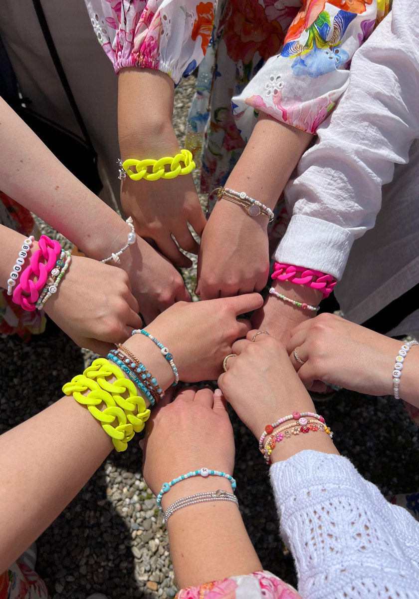 A group stands in a circle, extending hands adorned with Princess Goes Hollywood chunky Neon Pink or Neon Gelb bracelets—some crafted from recycled plastic and paired with colorful beads and floral-patterned sleeves over pebble-strewn ground.