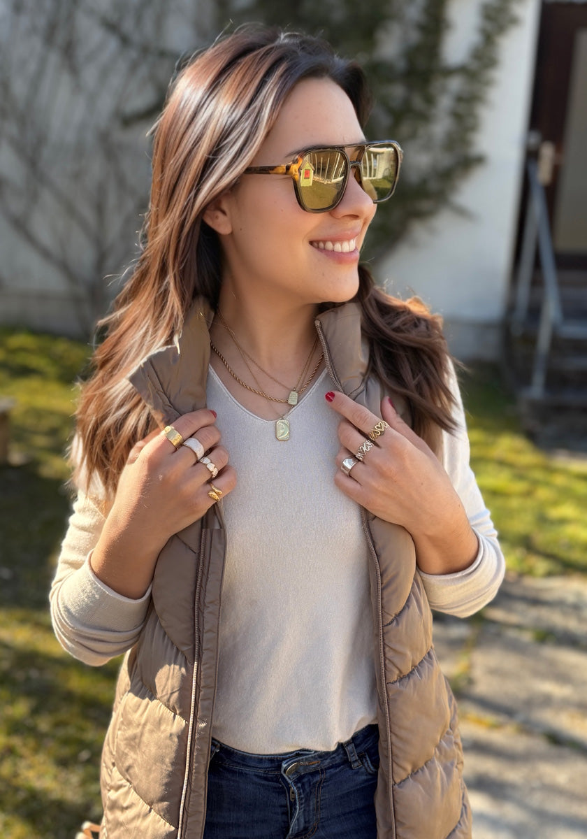A woman with long brown hair wears sunglasses, a beige puffy vest, and layered necklaces. She smiles outdoors, showing off her Princess Goes Hollywood Ring mit Herzen in Perlmutt-Optik.