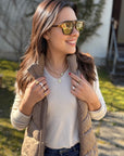 A woman outdoors smiles in tortoiseshell sunglasses, a beige quilted vest, and blue jeans. She wears layered necklaces, the Princess Goes Hollywood Ring in Perlmutt-Optik, and red nail polish amid sunlit greenery.