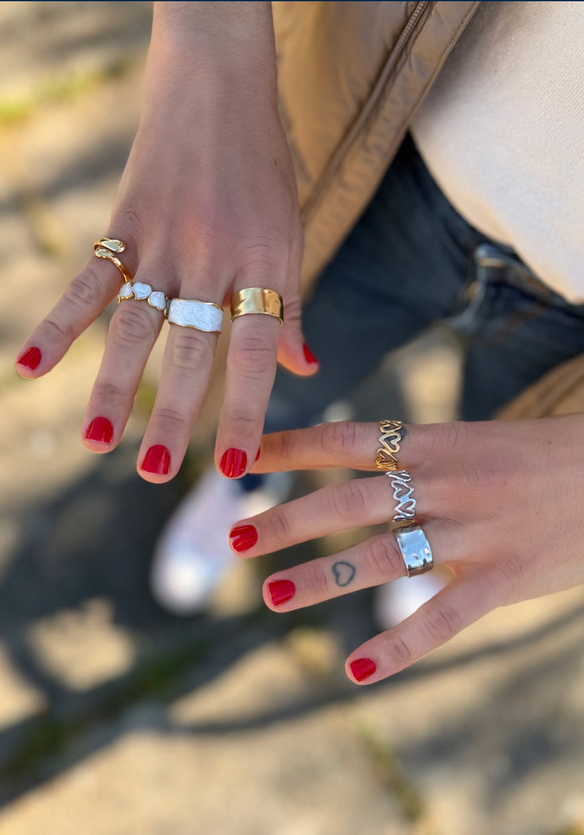 A person with red polish showcases both hands wearing gold and silver rings, including the Princess Goes Hollywood Ring mit Herzen in Perlmutt-Optik. One finger has a small green heart tattoo. Sunlight, jeans, a white shirt, and a beige jacket blur in the background.