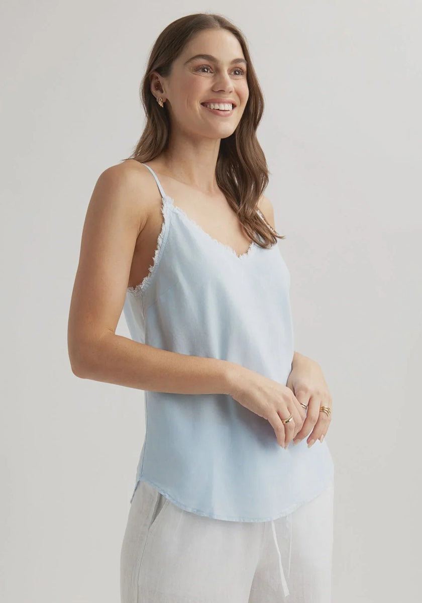 A woman with long brown hair smiles while wearing a light blue Bella Dahl Lyocell Trägertop with frayed edges and light pants, standing against a plain, light background, facing left with her hands loosely clasped.
