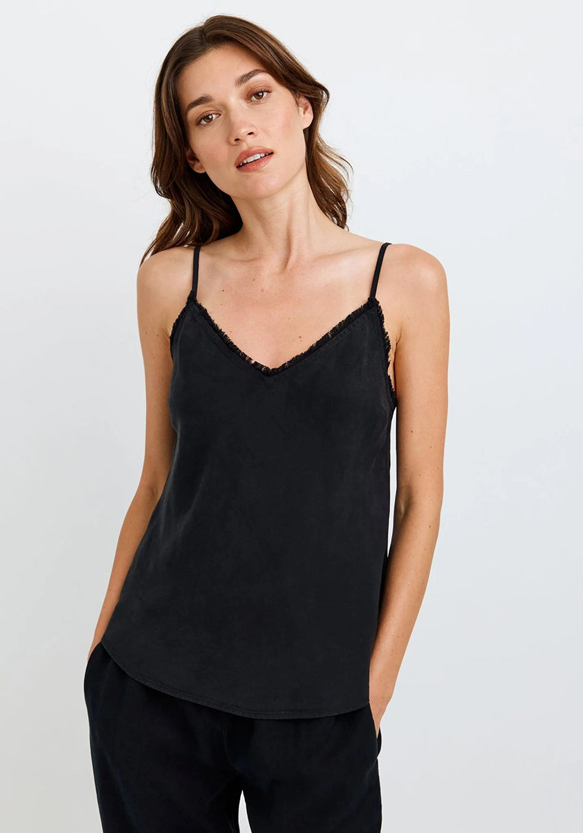 A woman with long brown hair wears a black Bella Dahl Lyocell tank top with thin straps and lace trim, paired with loose black pants. She stands against a white background, hands in pockets, gazing to the side with a neutral expression.