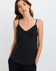 A woman with long brown hair wears a black Bella Dahl Lyocell tank top with thin straps and lace trim, paired with loose black pants. She stands against a white background, hands in pockets, gazing to the side with a neutral expression.