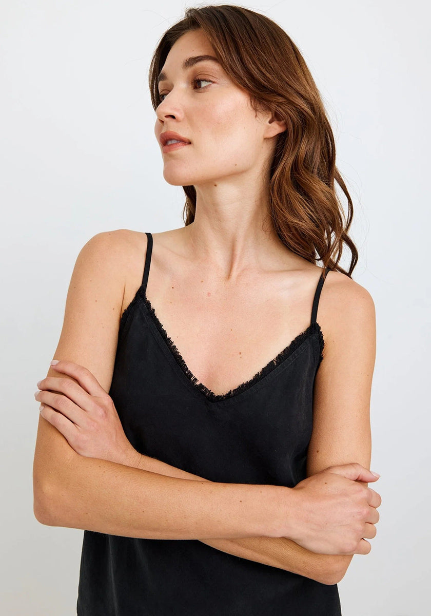 A woman with wavy brown hair stands against a plain light background, wearing a black Bella Dahl Lyocell Trägertop with a subtle frayed trim on the neckline. She looks to her right with a neutral expression, arms loosely crossed.