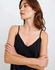 A woman with wavy brown hair stands against a plain light background, wearing a black Bella Dahl Lyocell Trägertop with a subtle frayed trim on the neckline. She looks to her right with a neutral expression, arms loosely crossed.