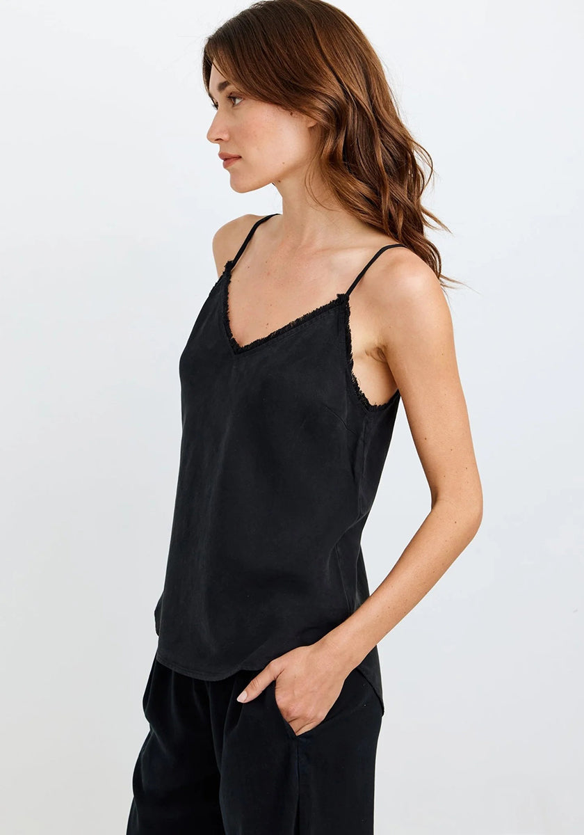 A woman with long, wavy brown hair stands in profile against a white background, wearing a Bella Dahl black lace-trim Trägertop and matching black Denimoptik pants, one hand in her pocket as she gazes ahead neutrally.