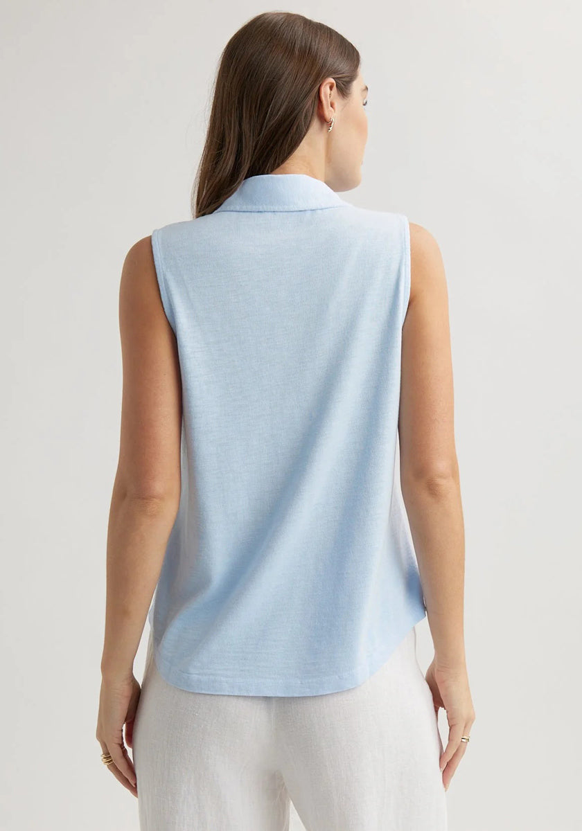 A woman with long brown hair is seen from the back, wearing a sleeveless, light blue Bella Dahl cotton-blend top with a shirt collar and white pants, standing against a plain light background, her arms relaxed by her sides.