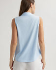 A woman with long brown hair is seen from the back, wearing a sleeveless, light blue Bella Dahl cotton-blend top with a shirt collar and white pants, standing against a plain light background, her arms relaxed by her sides.