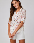 A woman with long brown hair smiles against a light background, wearing a Bella Dahl lightweight linen-blend blouse with floral print and white shorts. Her sleeves are rolled up, and her hands are loosely clasped in front.