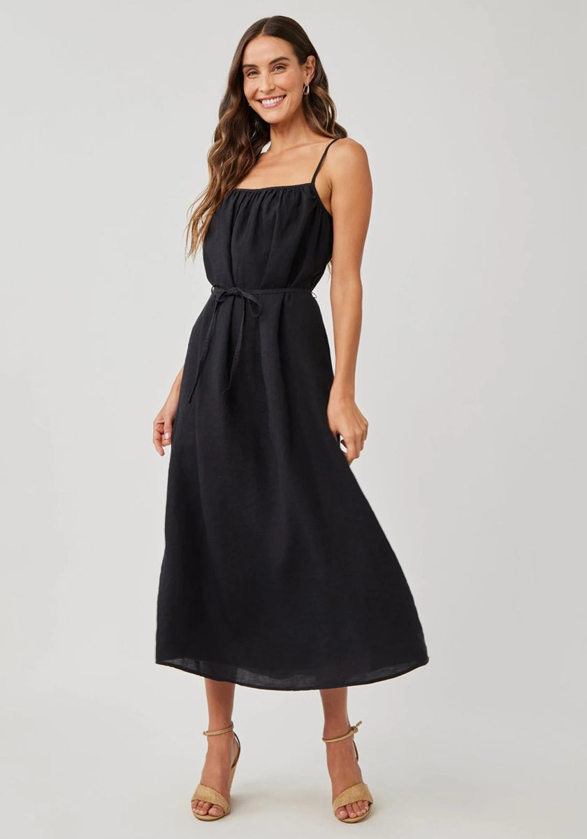 A woman with long wavy brown hair wears a Bella Dahl linen midi dress with adjustable straps, a tie belt, and thin stripes. She stands in light strappy heels, smiling with one hand in her pocket against a plain light background.