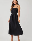 A woman with long wavy brown hair wears a Bella Dahl linen midi dress with adjustable straps, a tie belt, and thin stripes. She stands in light strappy heels, smiling with one hand in her pocket against a plain light background.