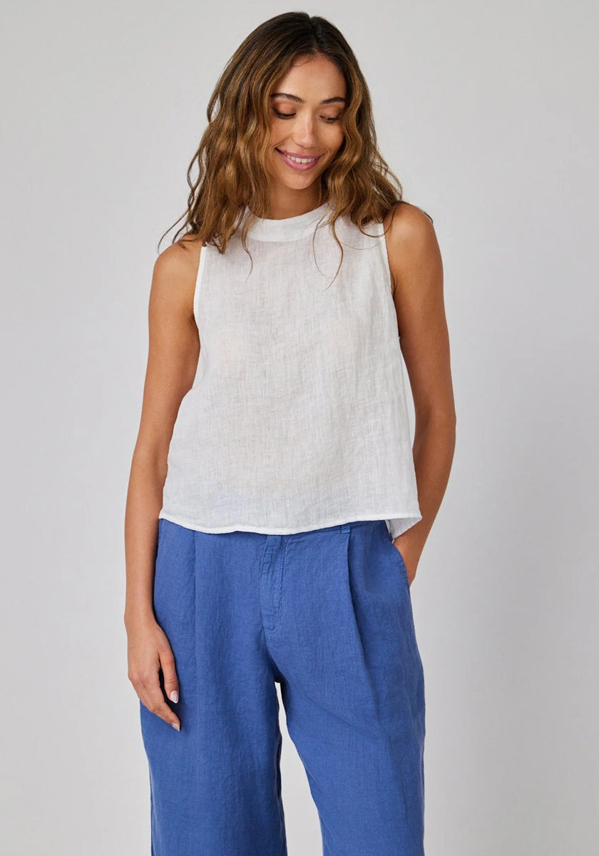 A woman with wavy light brown hair wears a sleeveless linen top with round neckline and tie details from Bella Dahl, paired with wide, high-waisted blue pants. She stands before a light gray background, smiling softly and looking down, one hand in her pocket.