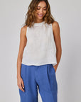 A woman with wavy light brown hair wears a sleeveless linen top with round neckline and tie details from Bella Dahl, paired with wide, high-waisted blue pants. She stands before a light gray background, smiling softly and looking down, one hand in her pocket.