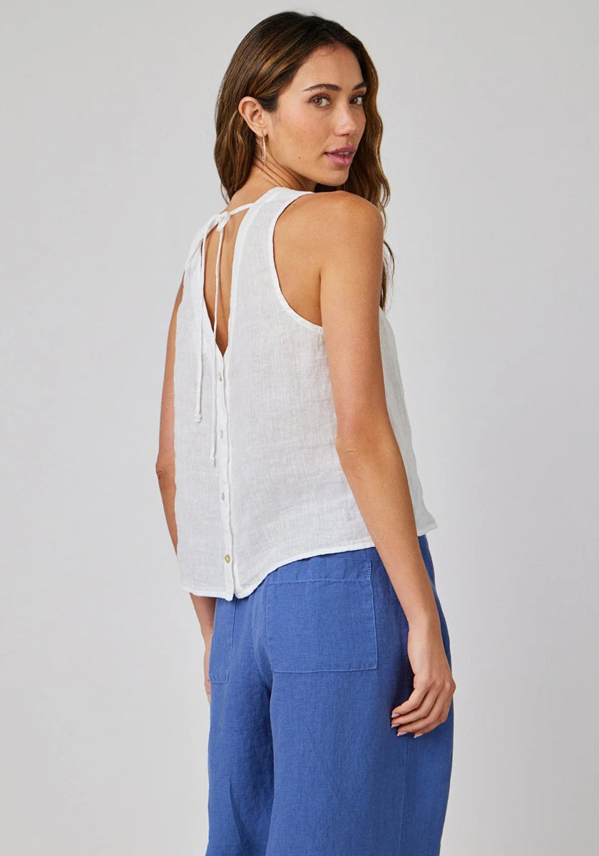 A woman with long, wavy hair stands sideways against a gray background, wearing a sleeveless Bella Dahl Leinen Top with round neckline, back buttons, and neck ties. She pairs it with loose-fitting, high-waisted blue pants.