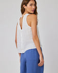 A woman with long, wavy hair stands sideways against a gray background, wearing a sleeveless Bella Dahl Leinen Top with round neckline, back buttons, and neck ties. She pairs it with loose-fitting, high-waisted blue pants.