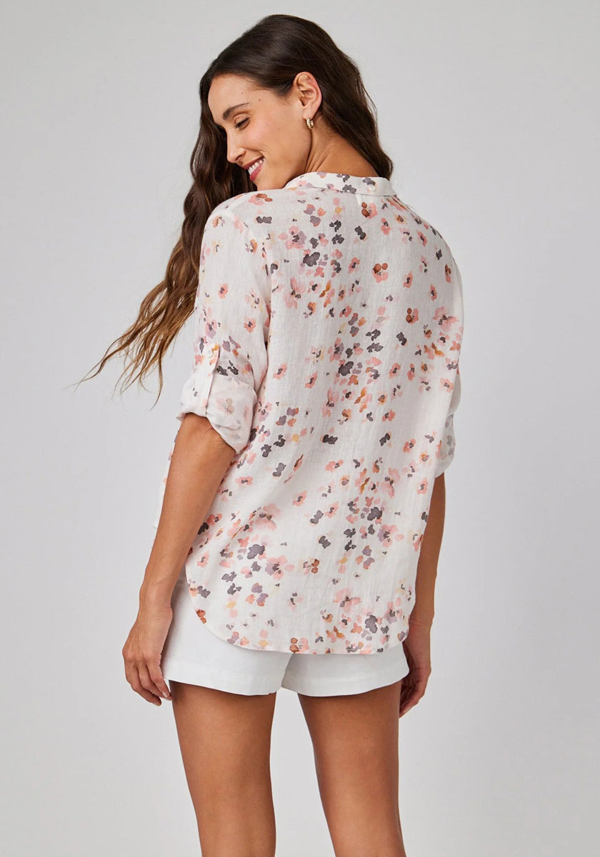 A woman with long, wavy brown hair stands facing away, smiling over her shoulder. She wears a light Bella Dahl Leinenmix Bluse mit Blumenprint and white shorts with rolled sleeves, set against a plain light gray background.